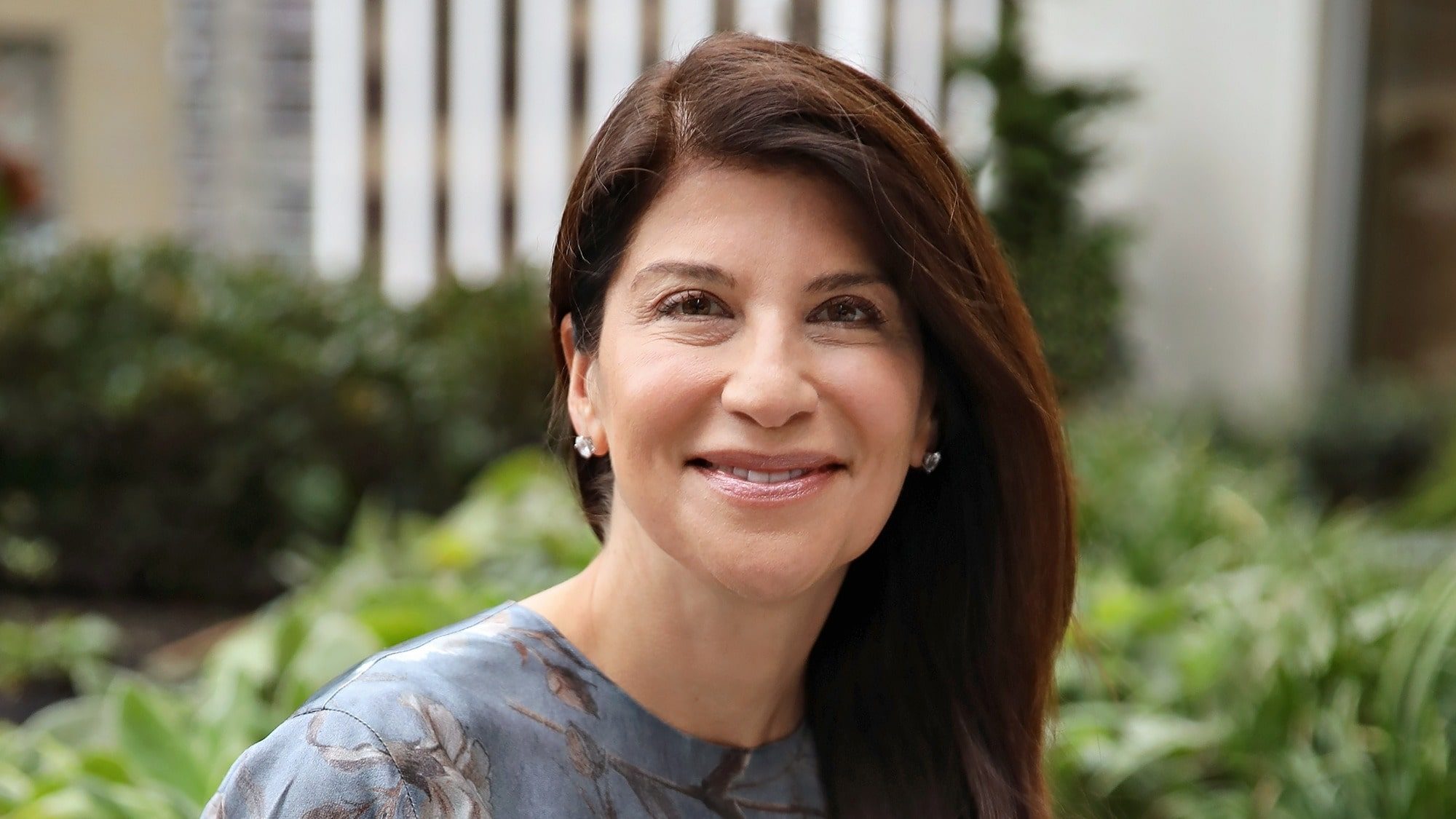 Portrait of a smiling woman with long brown hair, wearing a blue floral blouse, outdoors with blurred greenery and an investment catalyst building in the background.