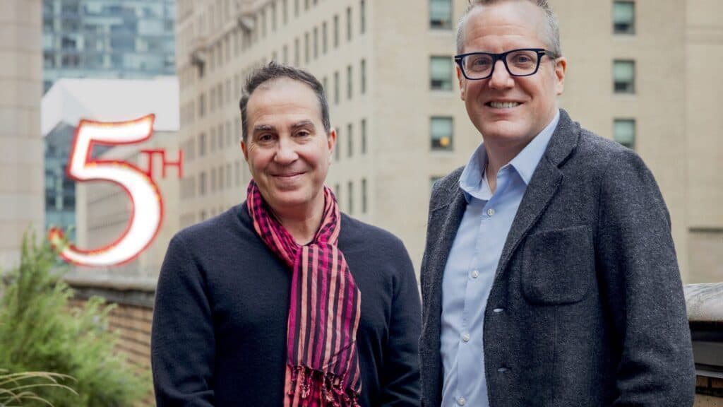 Two men stand outdoors in front of a building, with a large "5th" sign partially visible in the background.