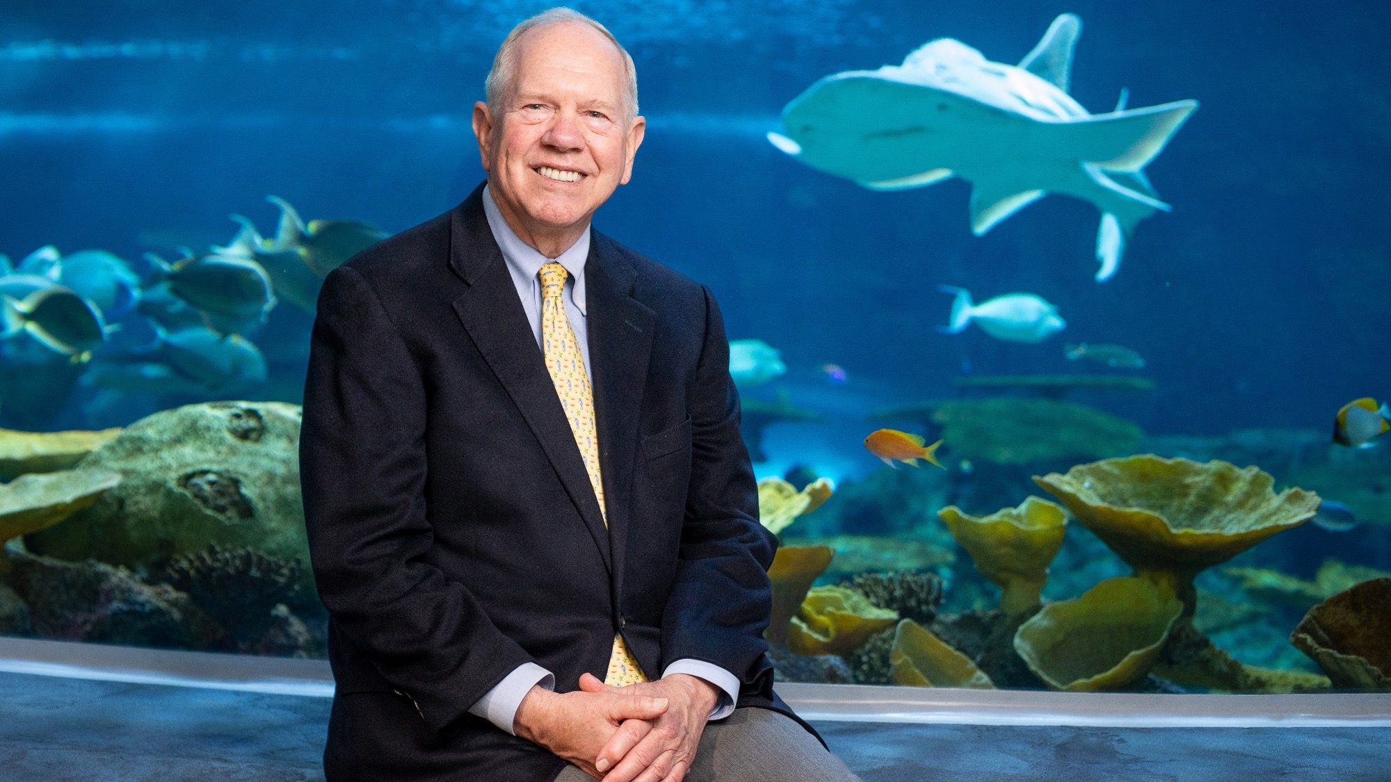 An elderly man in a suit, fitting the aura of a Sea Boss, sits in front of a large aquarium filled with various fish, including a prominent shark.