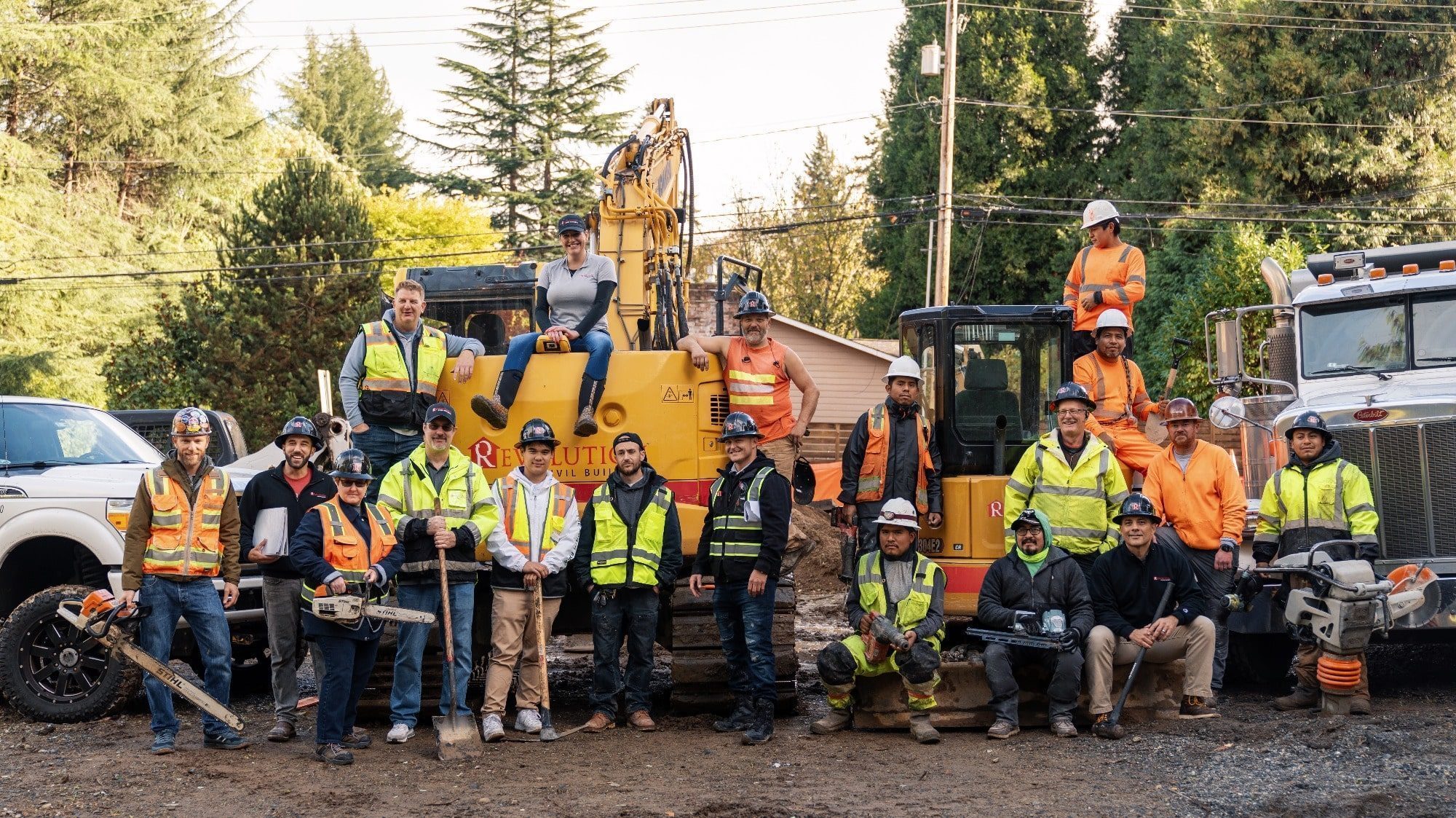 A group of construction workers wearing safety gear poses in front of construction machinery, including an excavator and a truck, amidst a wooded area reminiscent of the industrial spirit during the Cultural Revolution.