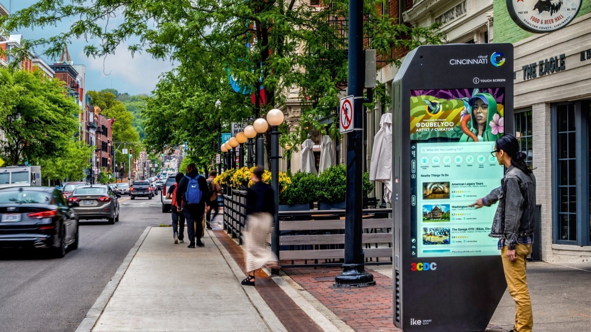 A person uses a large interactive touchscreen kiosk on a city sidewalk, with pedestrians and cars visible along the street.