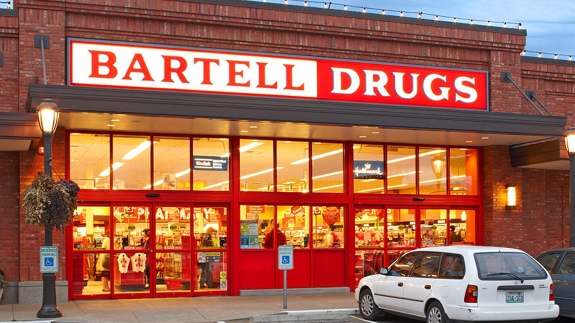 Exterior view of a Bartell Drugs store with bright red signage, large front windows, and a parked white car in front.
