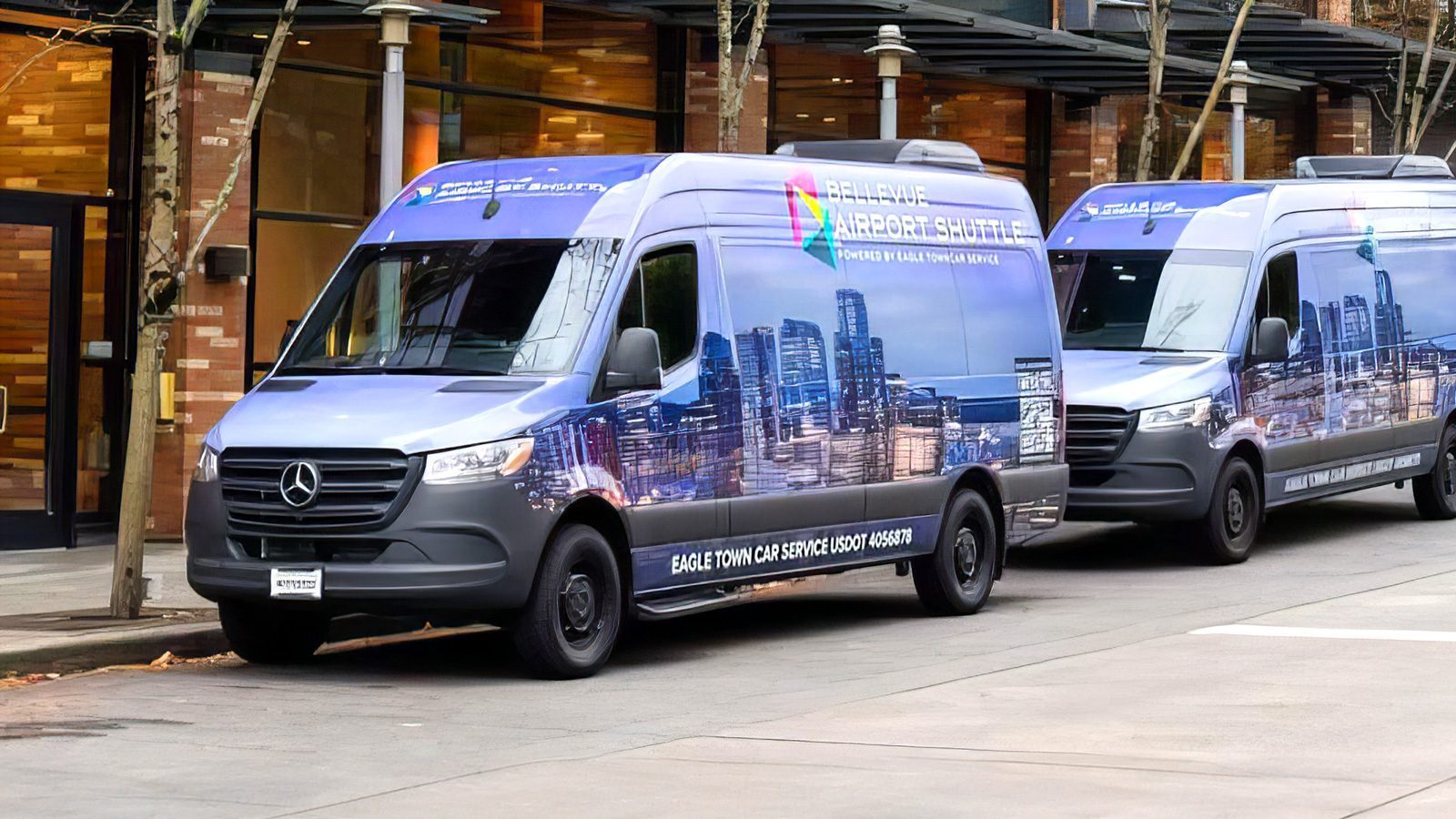 Two Mercedes-Benz Sprinter vans with cityscape advertisements are parked along a street with brick buildings and barren trees in the background.