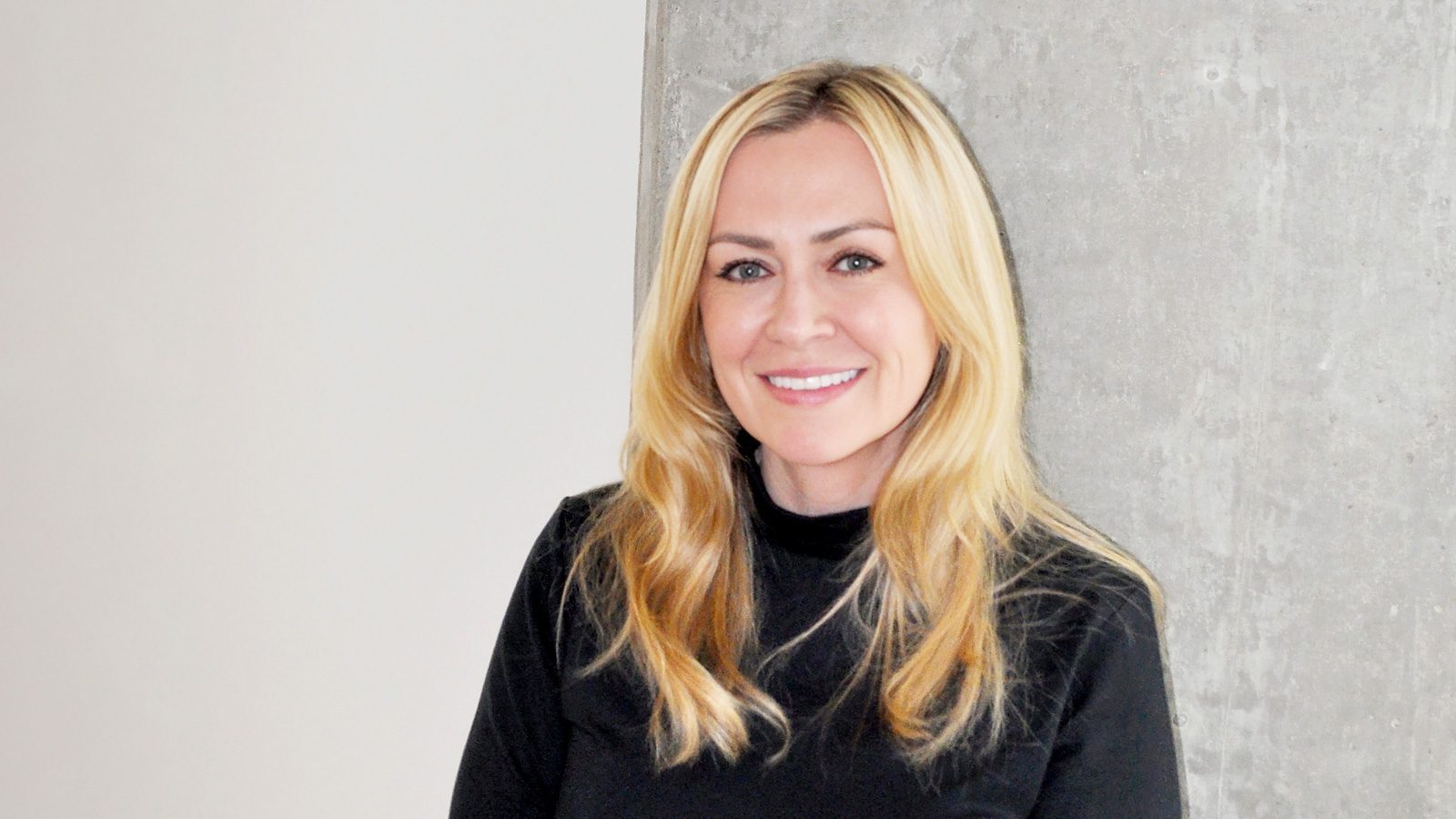 A person with long blonde hair is smiling while standing in front of a concrete wall, wearing a black top, embodying the spirit of employees' empowerment.