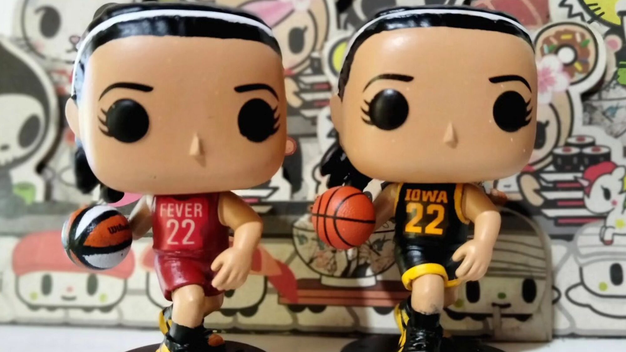Two Funko Pop! figures of female basketball players holding basketballs, one in a red Fever #22 uniform and the other in a black Iowa #22 uniform, stand side by side.