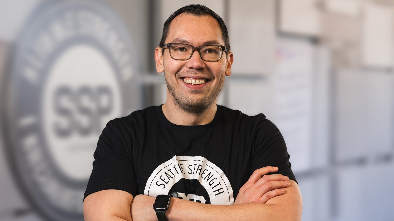 A person with glasses smiling and standing with arms crossed, wearing a black T-shirt with "Seattle Strength SSP" written on it, exemplifying well-being. The background features a blurred logo and some indistinct shapes.