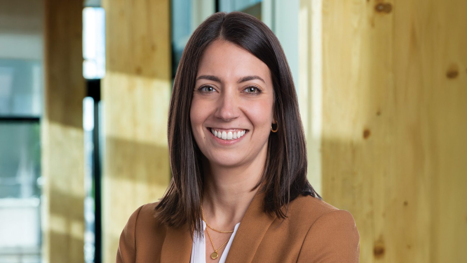 Woman with long brown hair, wearing a brown blazer and white shirt, smiles confidently in an office setting, celebrating the success of landing a job after college.