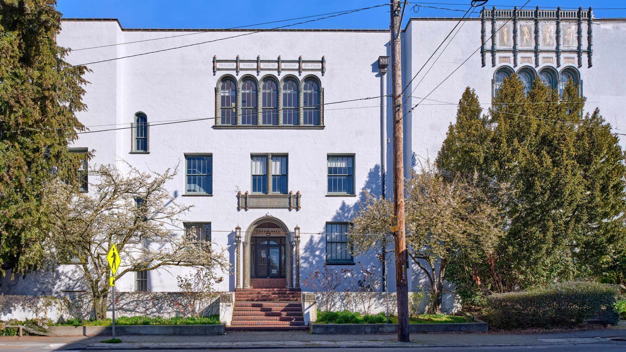 A white stucco building, known as Kerry Hall for sale, with arched windows and a wooden door, flanked by trees, under a clear blue sky.