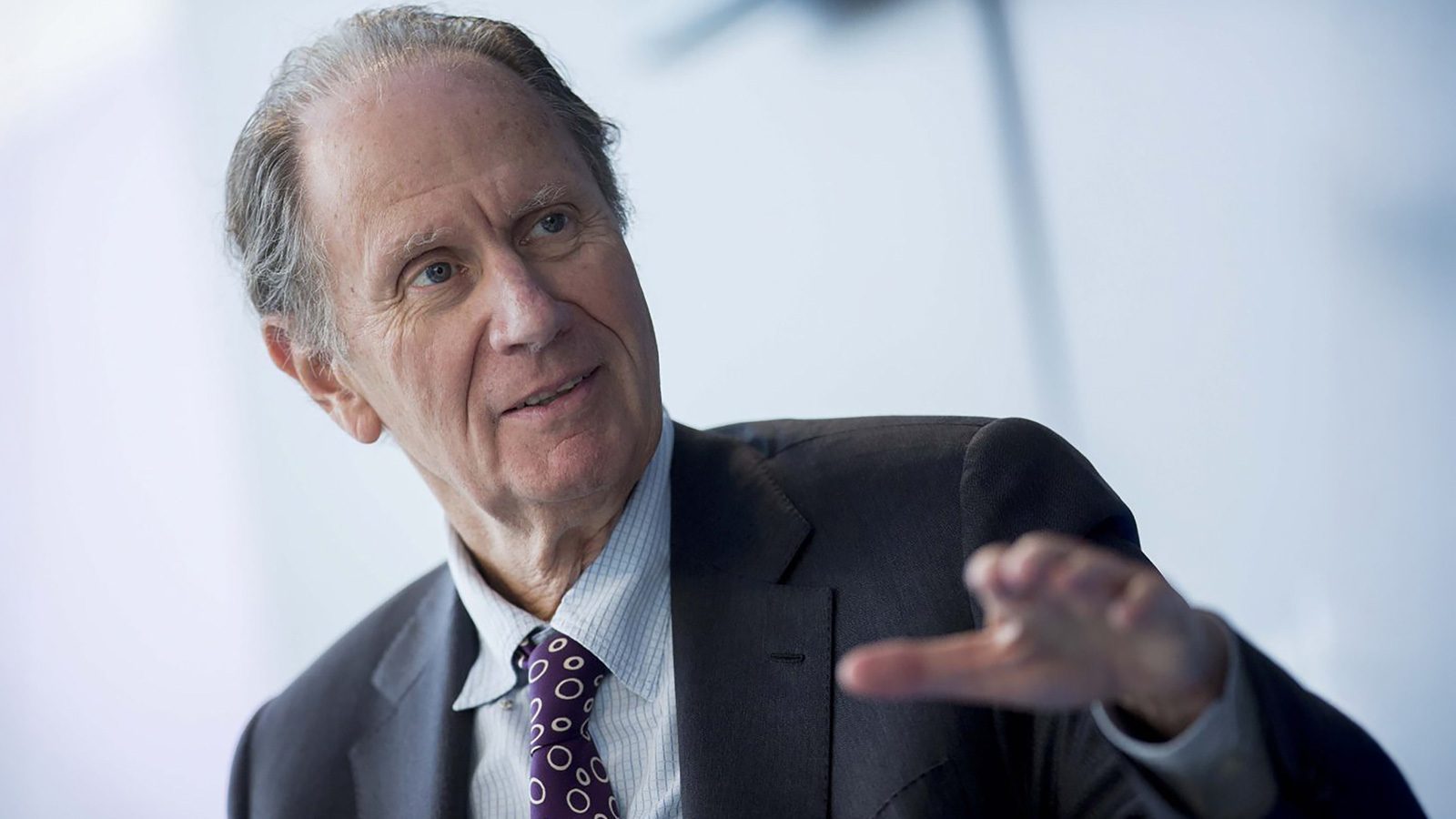 An older man in a suit, reminiscent of influential figures like David Bonderman, gestures while sitting in an office setting with a blurred backdrop.