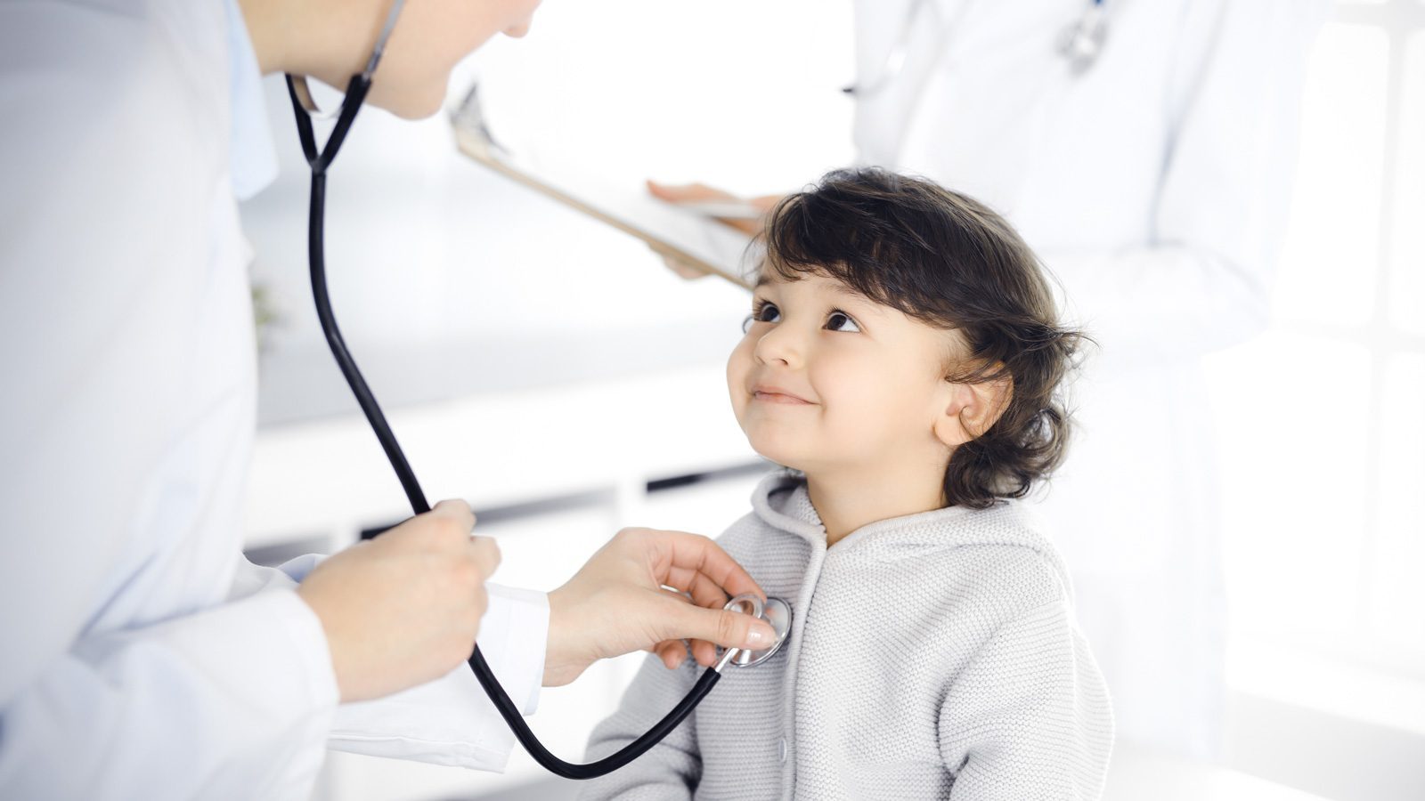 A doctor uses a stethoscope to examine a smiling young child in a clinic, showcasing kid-friendly design. Another medical professional holding a clipboard is visible in the background.