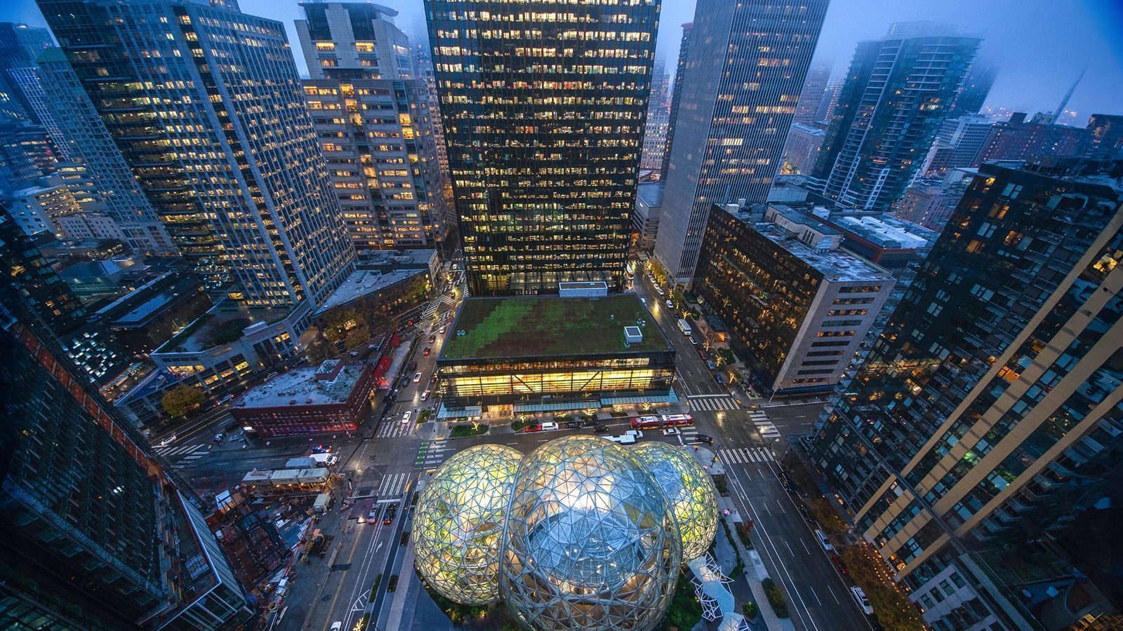 Aerial view of an urban cityscape at dusk, showcasing Seattle's influence with tall buildings surrounding a glass-domed structure filled with greenery and bustling city streets with traffic below.
