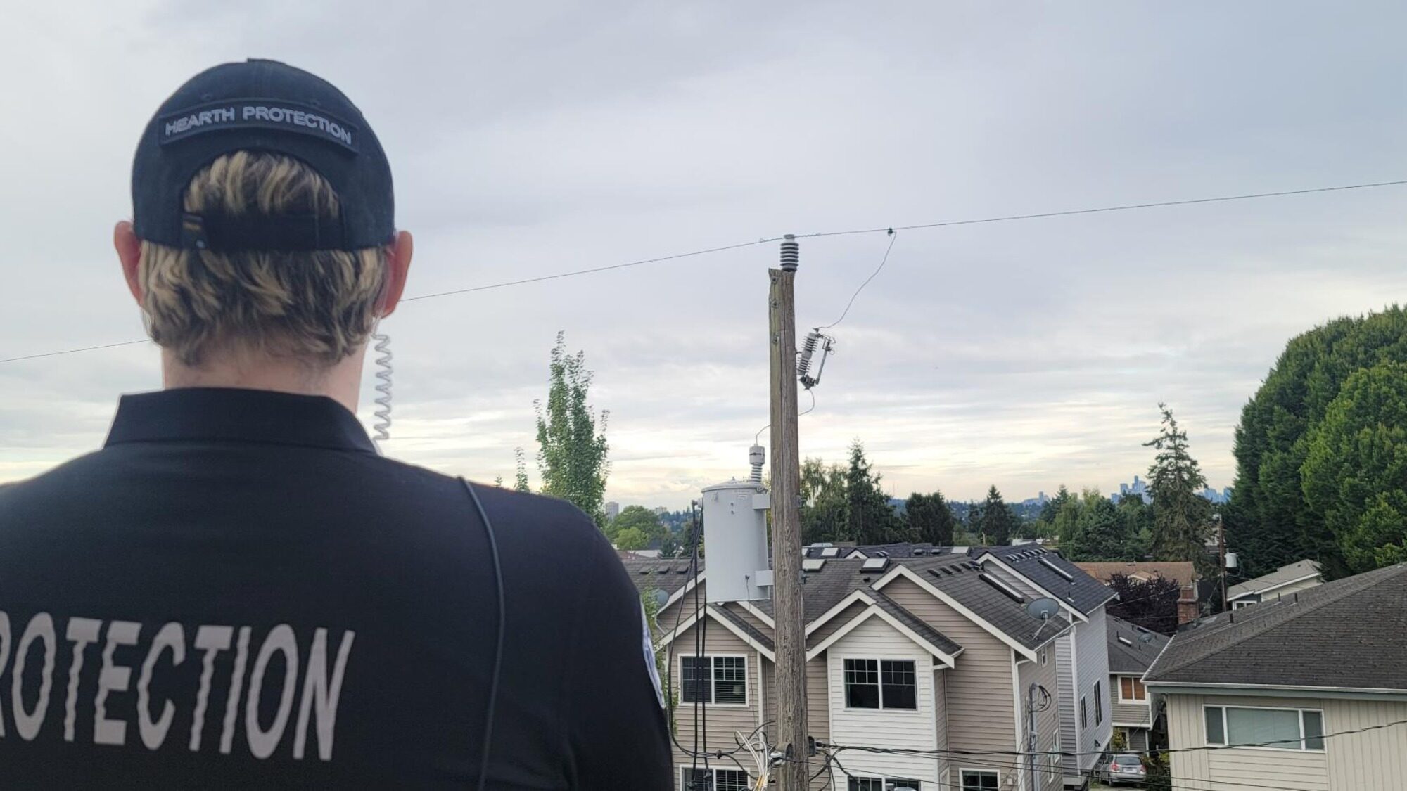 A person in a black "Protection" uniform and cap with a headset stands facing a residential neighborhood, embodying the new approach of Seattle's security industry amidst the trees and houses under a cloudy sky.
