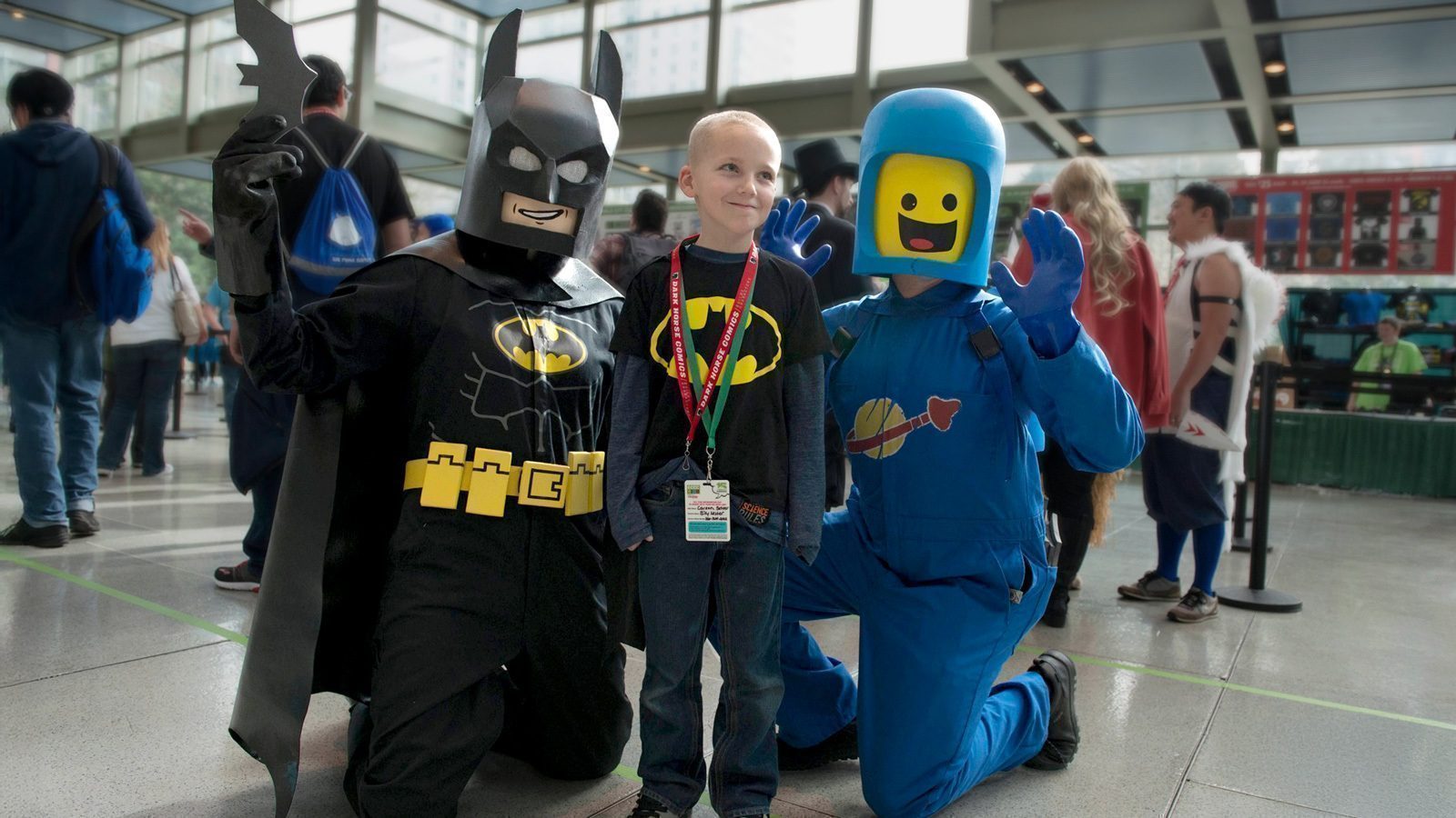 A child poses with two people in Batman and LEGO astronaut costumes at a convention center.