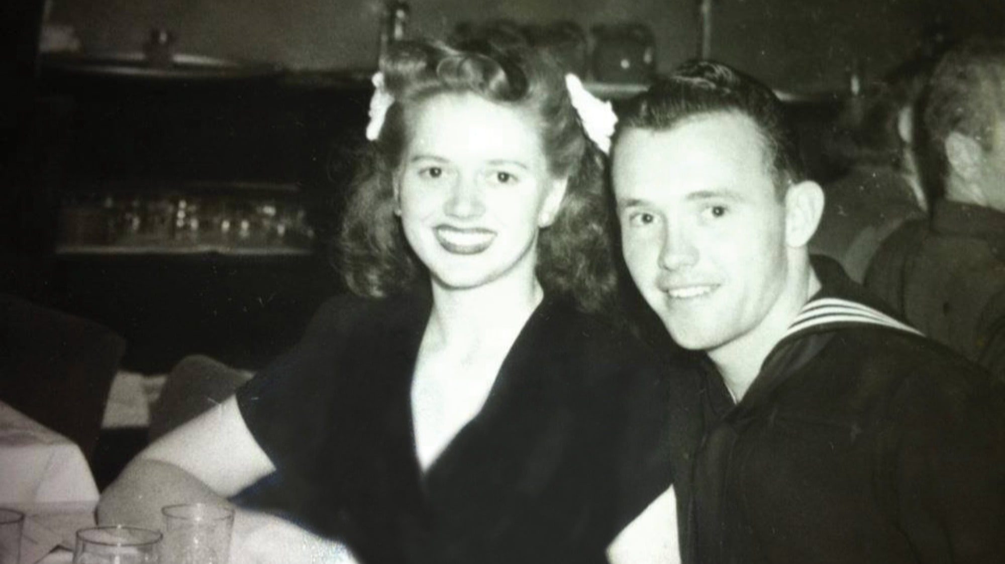A smiling woman and man dressed in a sailor uniform sit together at a table in a black-and-white photograph, reminiscent of the golden days, highlighting the timeless bond often cherished in elderly care.