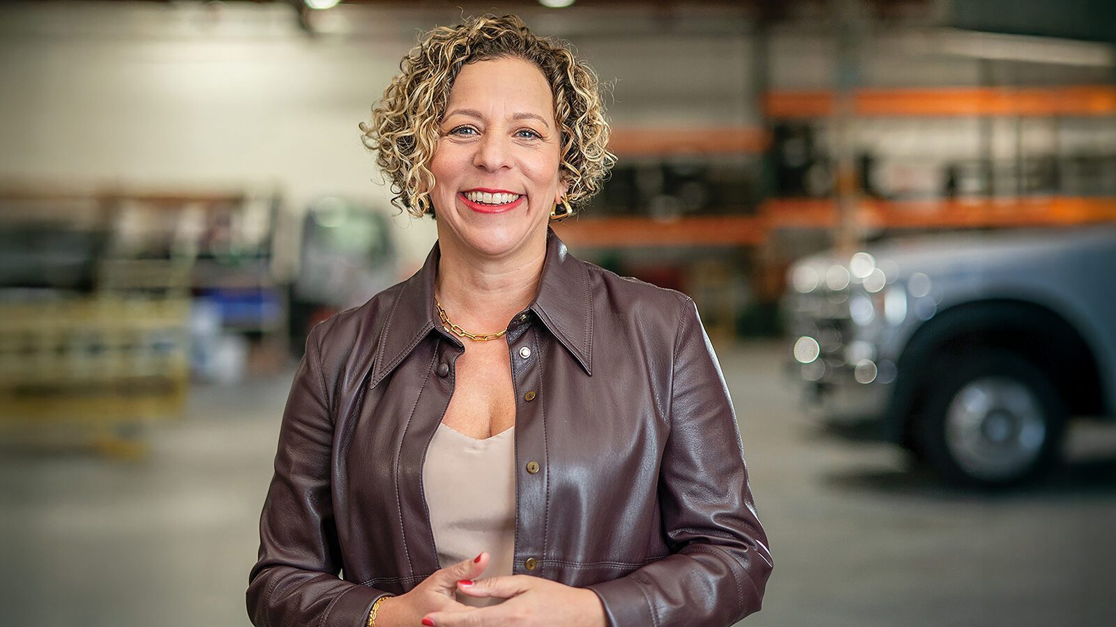Heather Snavely, a woman with short curly hair in a brown leather jacket, stands smiling in a warehouse with vehicles and shelves in the background, showcasing her role at AAA.