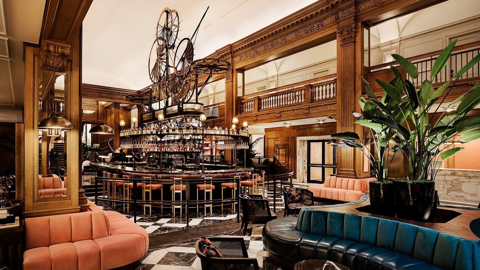 Elegant bar interior with a central circular bar, plush seating, and ornate wooden decor reminiscent of the historic Fairmont Olympic in Los Angeles. Large sculptures hang above, and plants add greenery to the space.