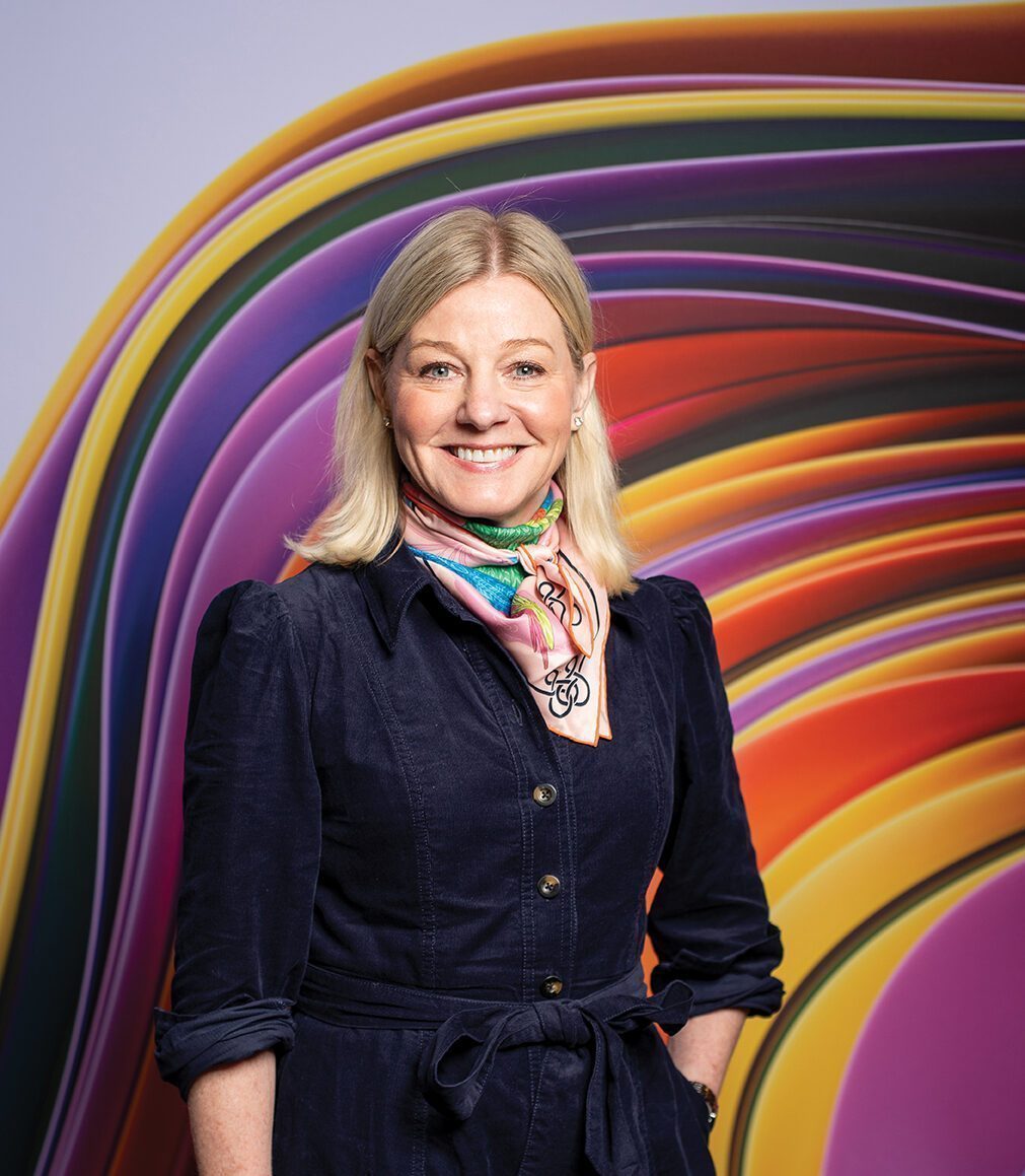 A woman with blonde hair, in a dark dress and colorful scarf, stands before a vibrant abstract background, evoking the dynamic growth fueled by tech companies investing in Seattle.