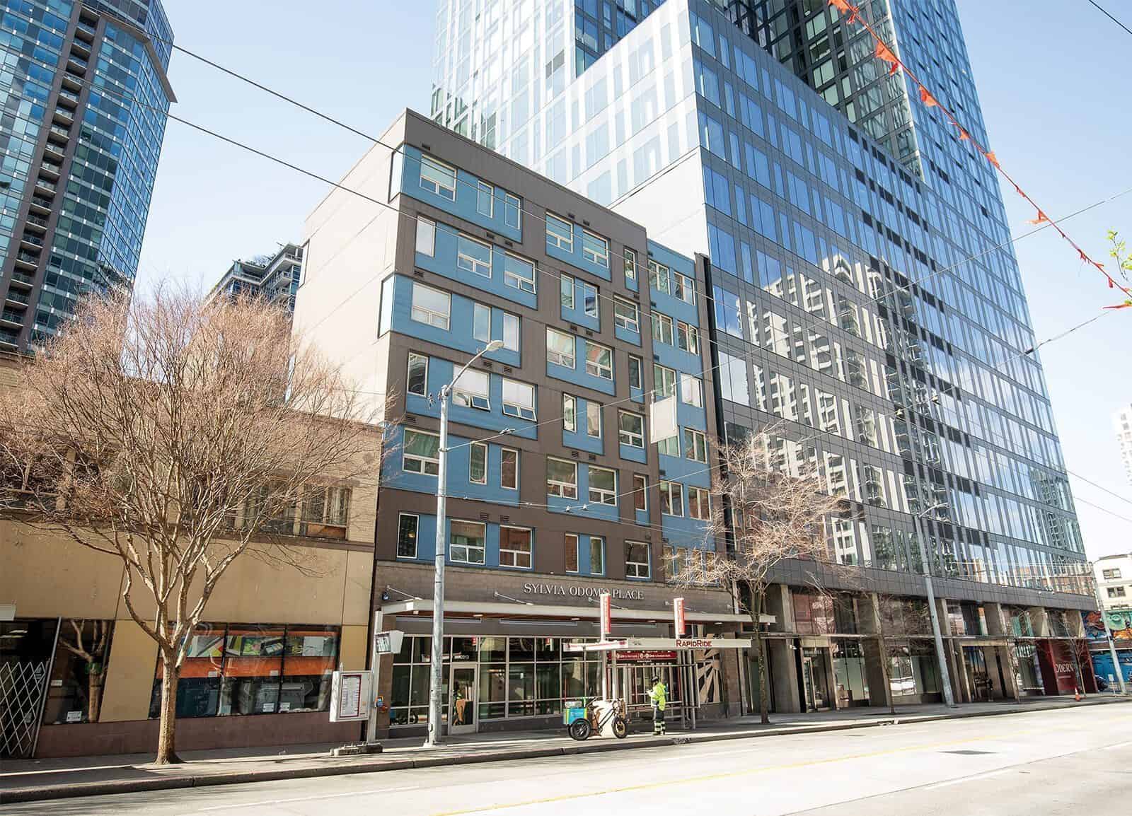 A mid-rise building with large windows and a ground floor awning stands on a city street, flanked by taller glass buildings and leafless trees—a reflection of tech companies investing in Seattle’s vibrant urban landscape.