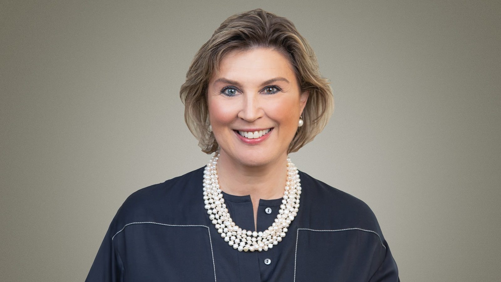 A woman with short, light brown hair wearing a dark blouse and a pearl necklace, smiling at the camera against a gray background, embodies the charm of the Land of Enchantment.