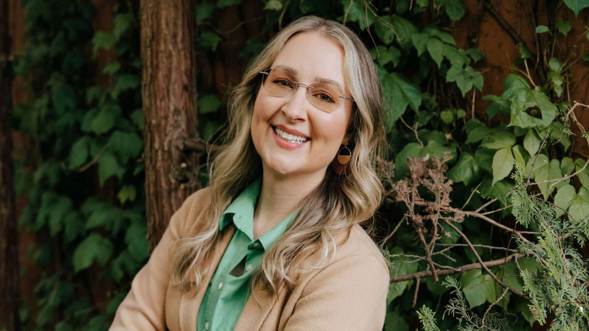 A person with long, wavy hair and glasses smiles while standing in front of a leafy and wooded background, embodying the joy and spontaneity of "Play's the Thing.
