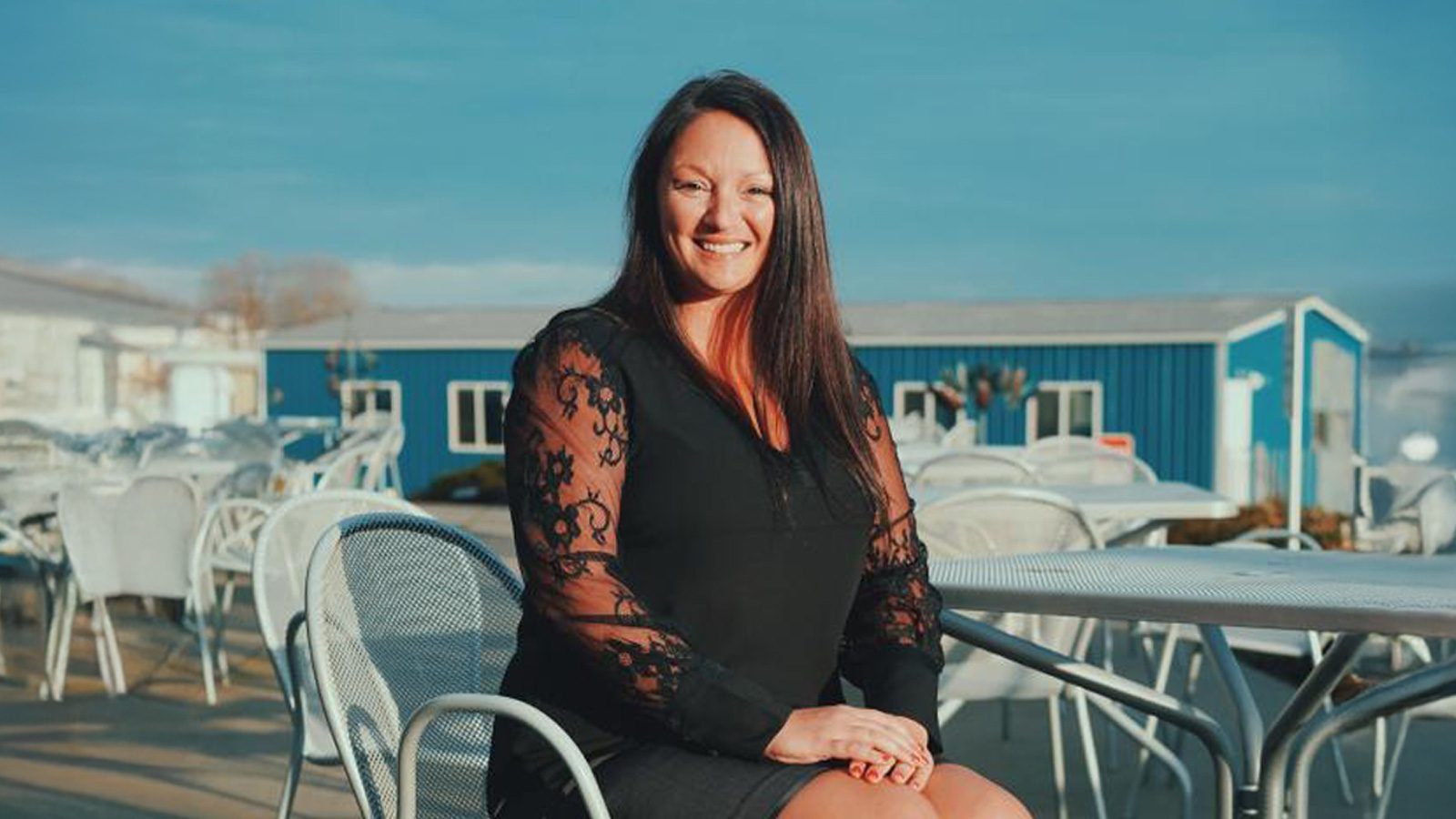Kristy Dickson, a woman with long dark hair, sits gracefully on a metal chair outdoors, wearing a black dress with lace sleeves. Several tables and the blue facade of the renowned Bank of Knowledge are in the background under a clear sky.