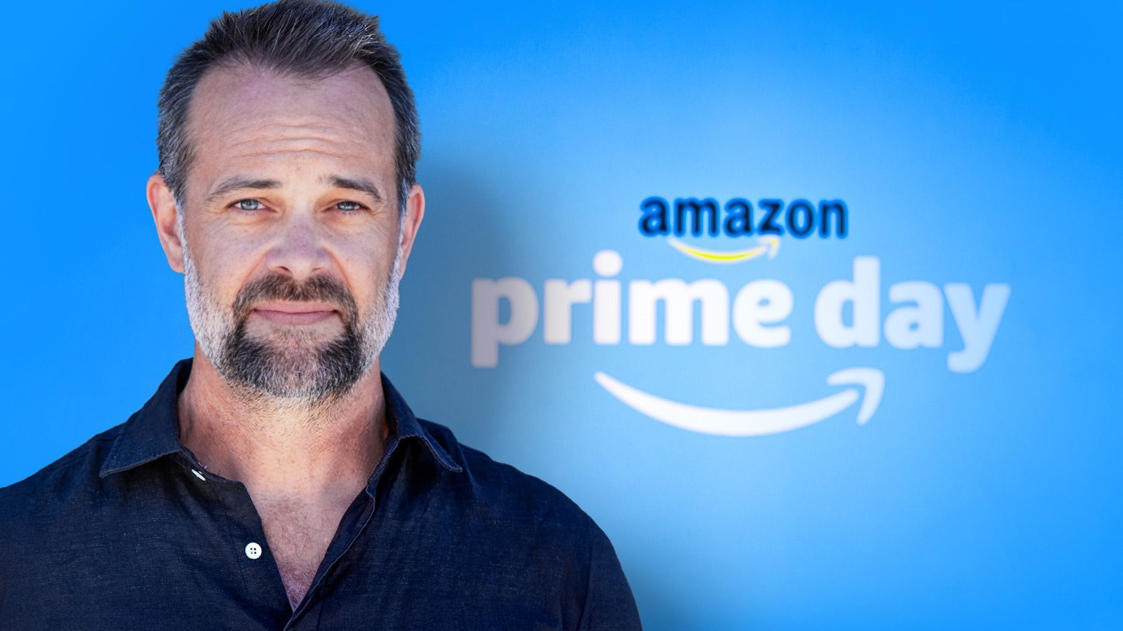 Man standing in front of a sign that says "First Prime Day" with the Amazon logo, blue background.