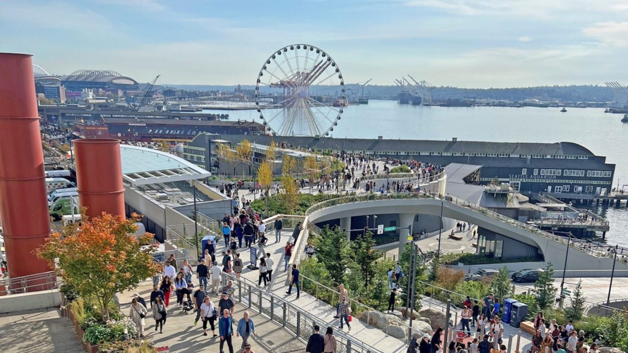An aerial view of Downtown Seattle's waterfront showcases a bustling area with a Ferris wheel, numerous people strolling along paths, and ships in the harbor under a clear sky.