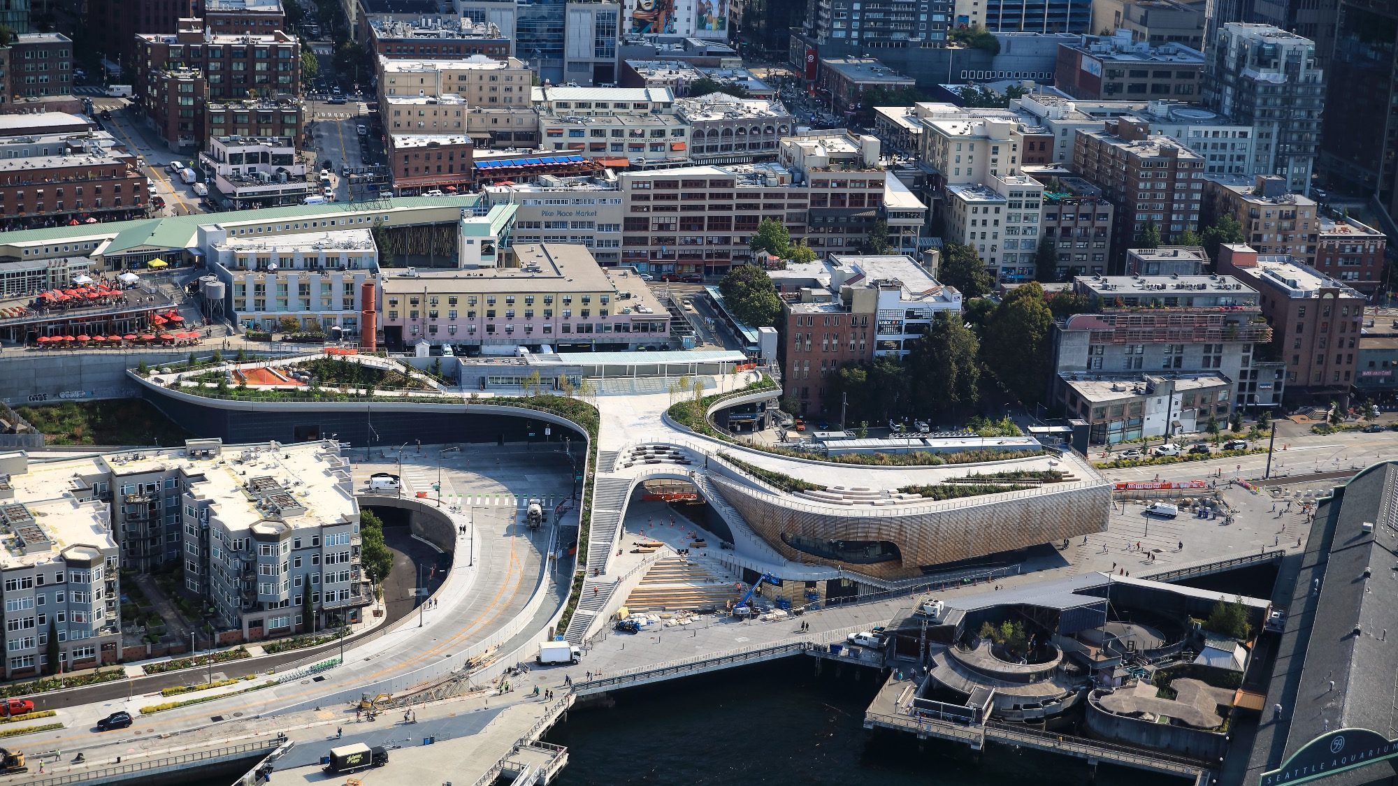 Aerial view of a modern urban landscape featuring the transformative Overlook Walk, a curved architectural structure gracefully integrated into the Seattle waterfront, surrounded by buildings and roads.