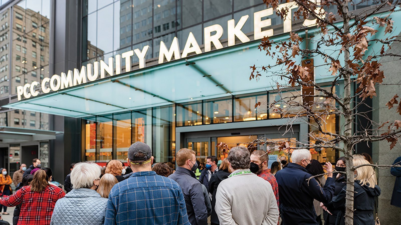 A crowd of people stands outside the entrance of PCC Community Markets in downtown Seattle, under an overhang with modern glass and metal architecture, eagerly anticipating the store reopening.