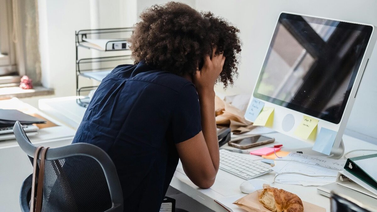 A person sits at a desk with their head in their hands in front of a computer, surrounded by papers, sticky notes, and a croissant, suggesting stress or frustration.