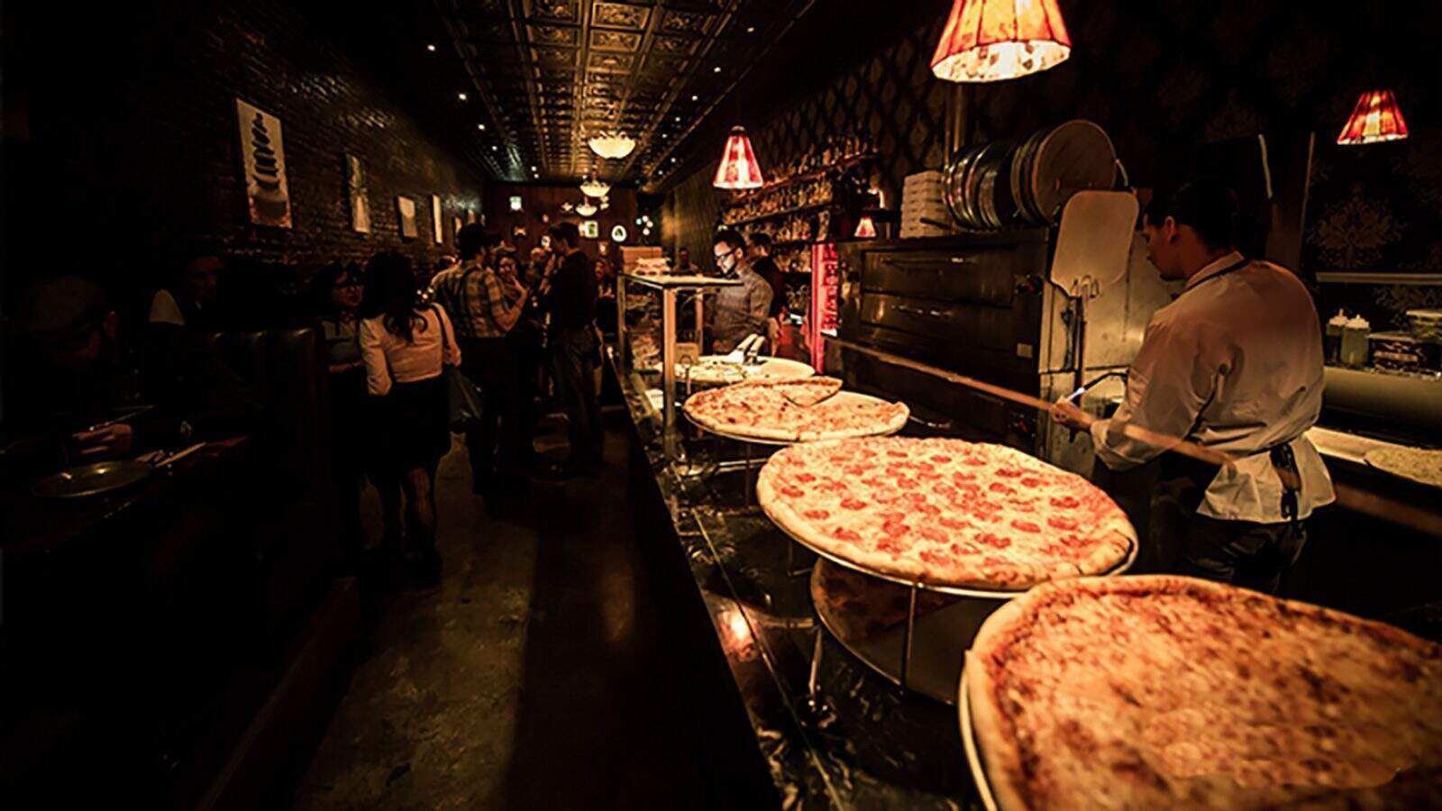 A dimly lit restaurant interior with large Seattle pizzas on a counter. A chef is preparing a saucy serenade of flavors, while patrons are seated and standing in the background.