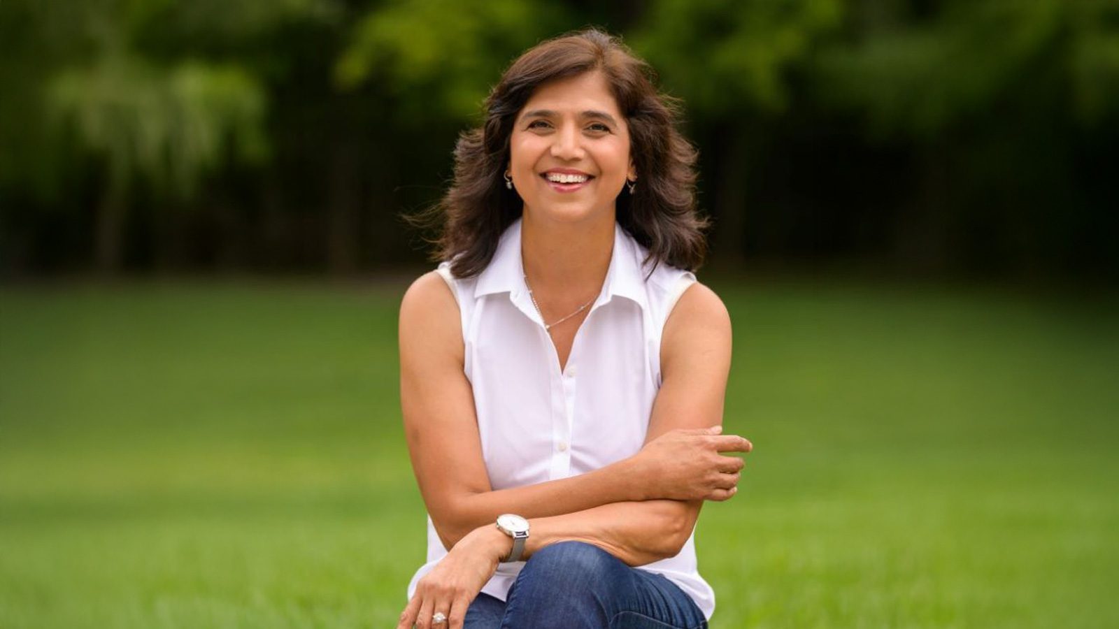 A person sitting outdoors on grass, wearing a white sleeveless top and jeans, with short dark hair, smiling and arms folded. Surrounded by lush greenery, they radiate confidence as if celebrating Big STEM Steps in their journey.