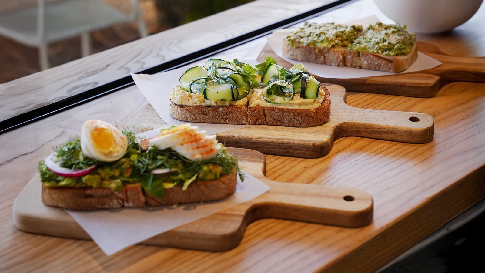 Three open-faced sandwiches with various toppings, including sliced egg, vegetables, and herbs, are arranged on wooden boards at Sano's Eastside locations.