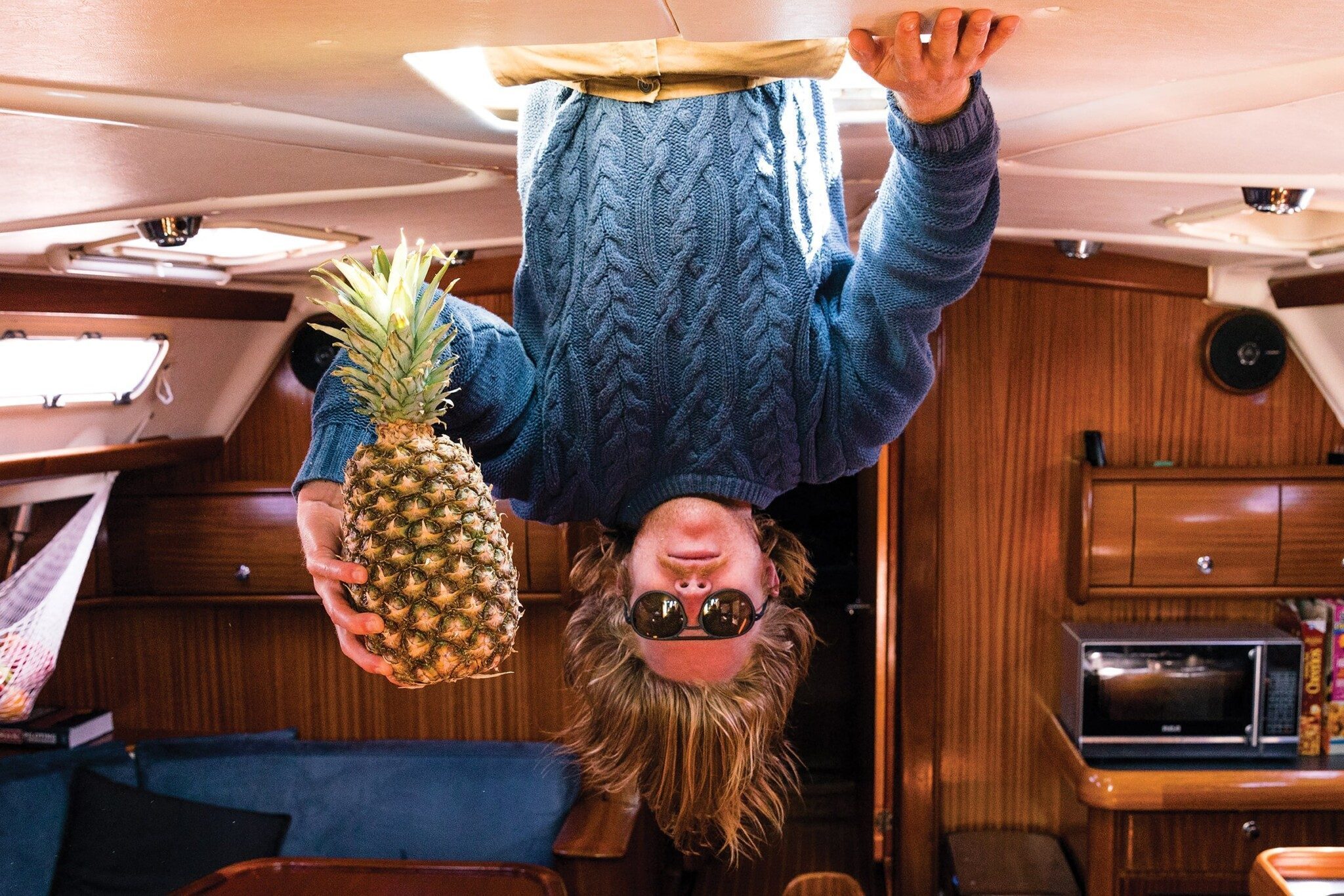 A person wearing sunglasses and a blue sweater holds a pineapple inside a wooden cabin. With quick thinking, they seem to transform the cabin into an exotic dessert wonderland. The image is intriguingly upside down.