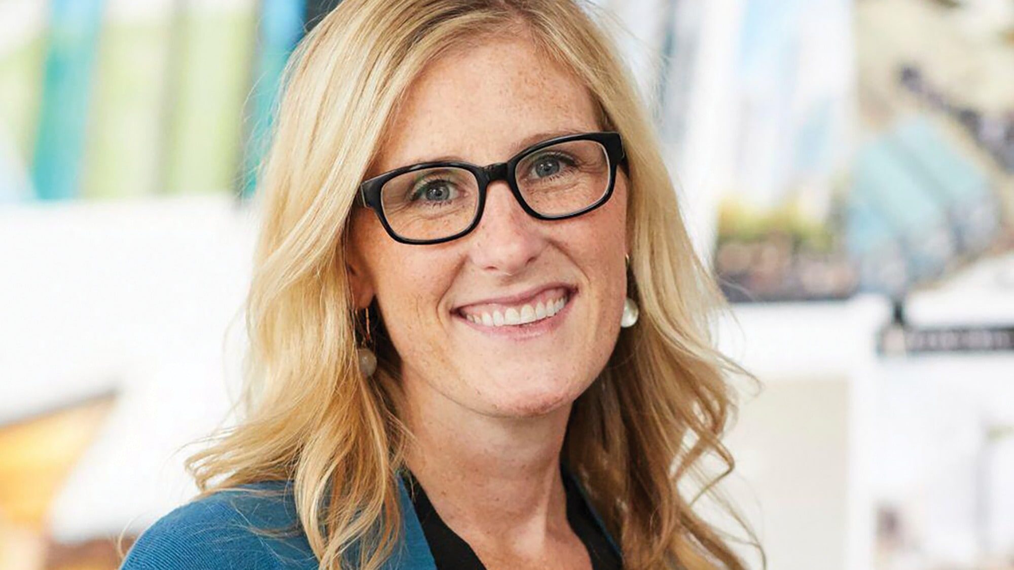 Mary Henson, captivated by architecture, smiles at the camera with long blonde hair and glasses, wearing a blue jacket. The background is blurred, drawing focus to her cheerful expression.