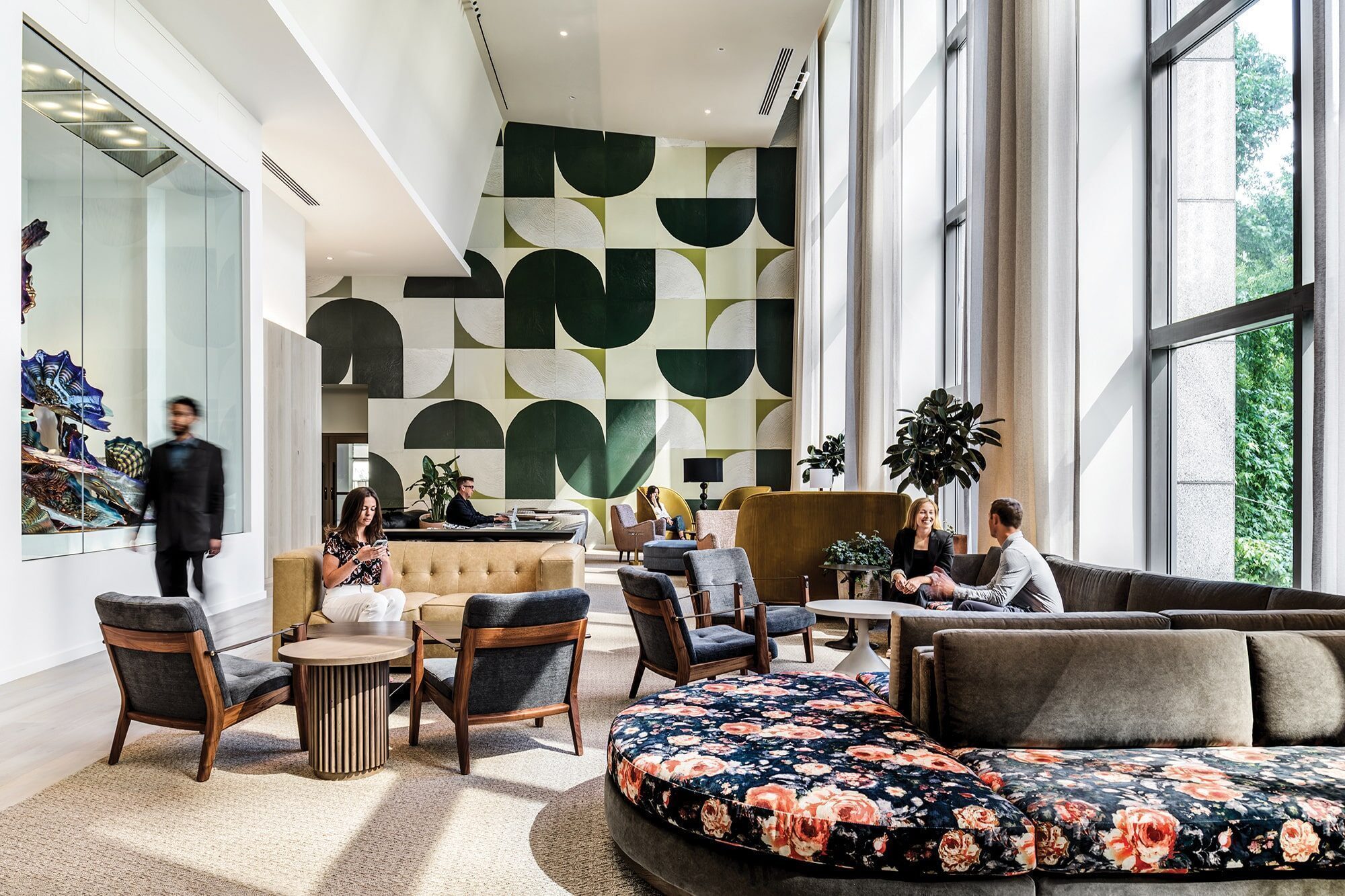 Modern lounge with abstract green wall art inspired by the career of architect Kyle Gaffney, featuring a mix of patterned and solid seating. Large windows flood the room with light as people sit, converse, and a person strolls on the left.