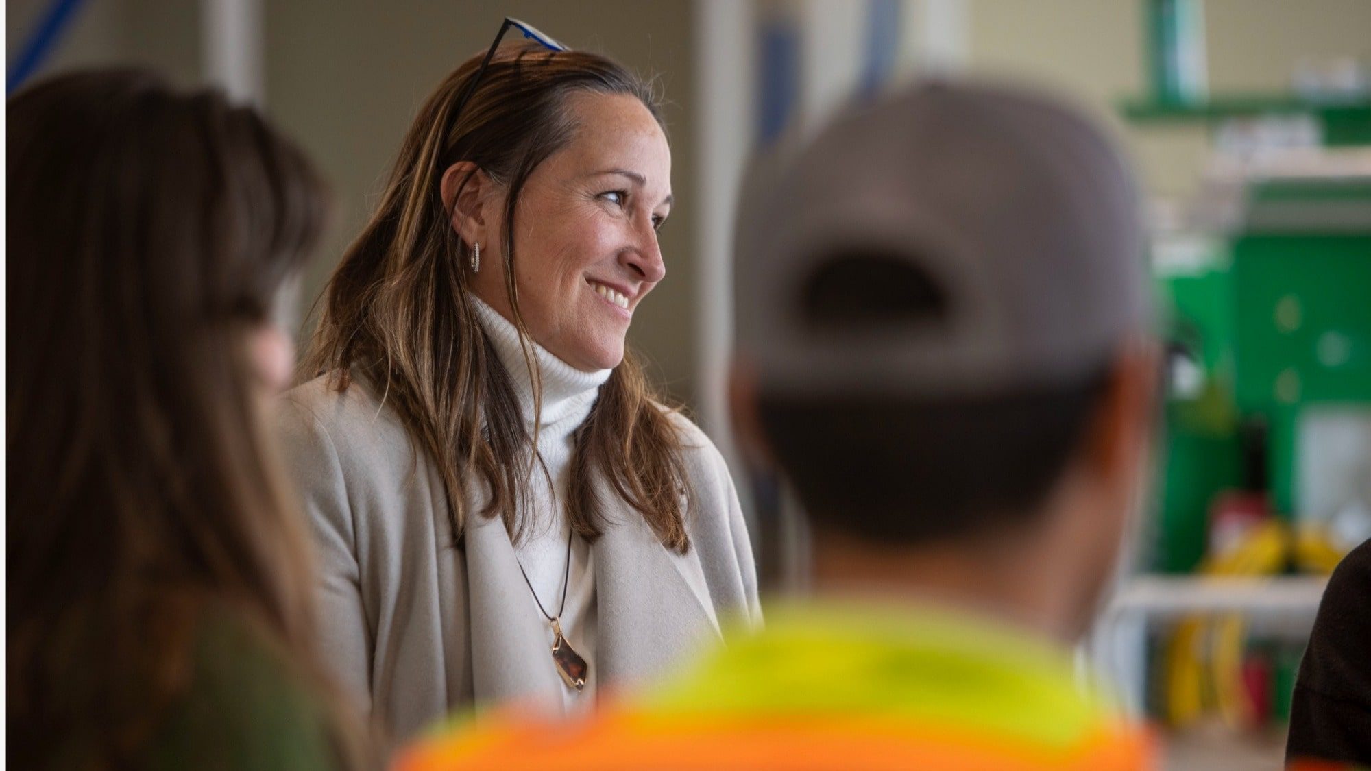 A woman wearing a white turtleneck and light-colored coat smiles while standing among other people in the room, her expression reflecting the hopeful beginnings of her medical journey.