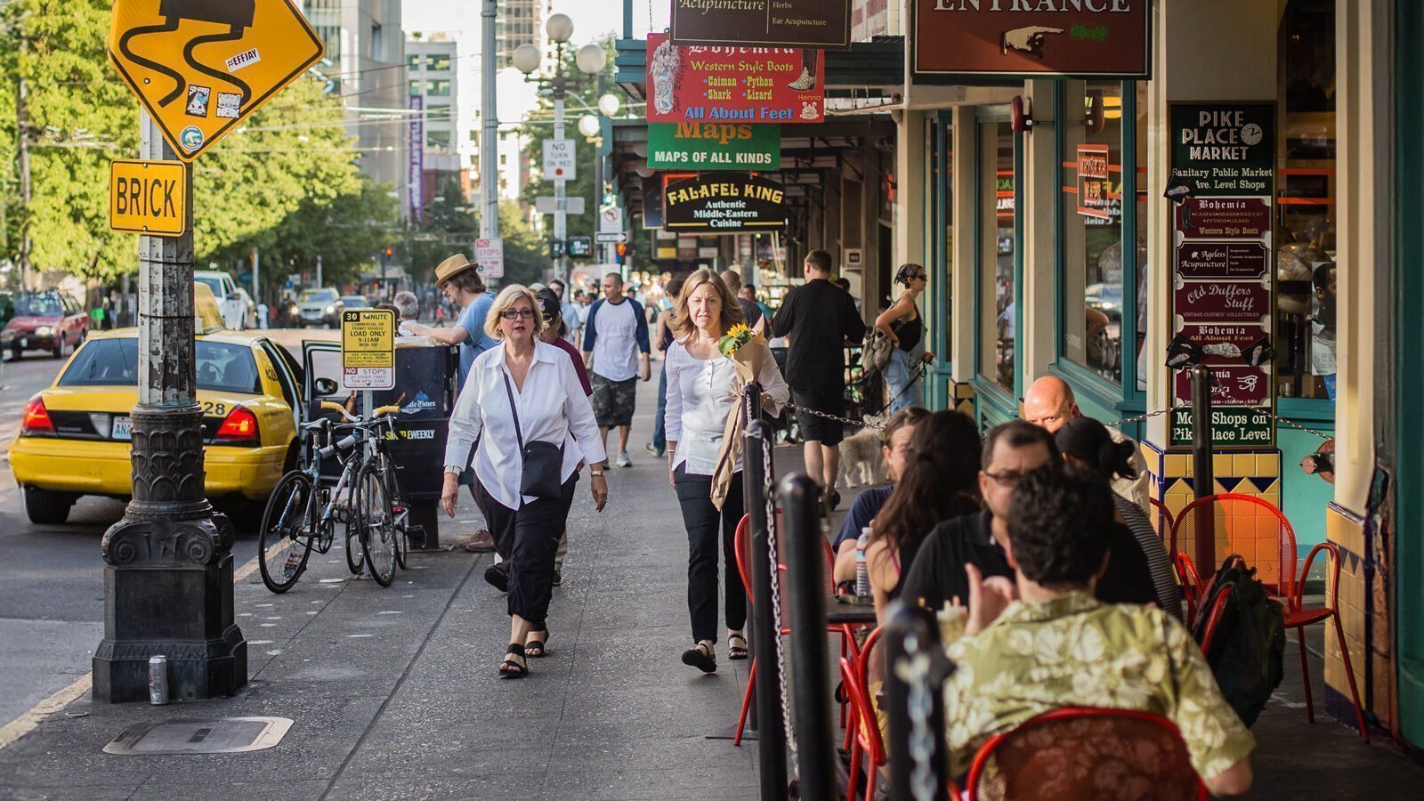 People walking on a busy sidewalk lined with shops and outdoor diners, near a street with cars and bicycles.