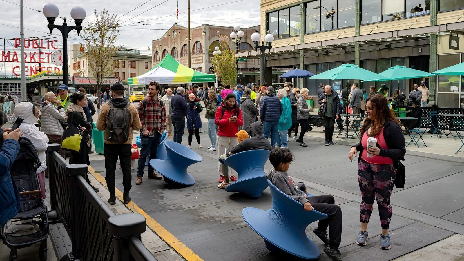 In this lively street scene, The Changing Face of First and Pike unfolds. People mingle at the market, some perched on blue spinning chairs while others exchange pleasantries. Behind them, a building adorned with green umbrellas stands as a backdrop to the vibrant interactions.