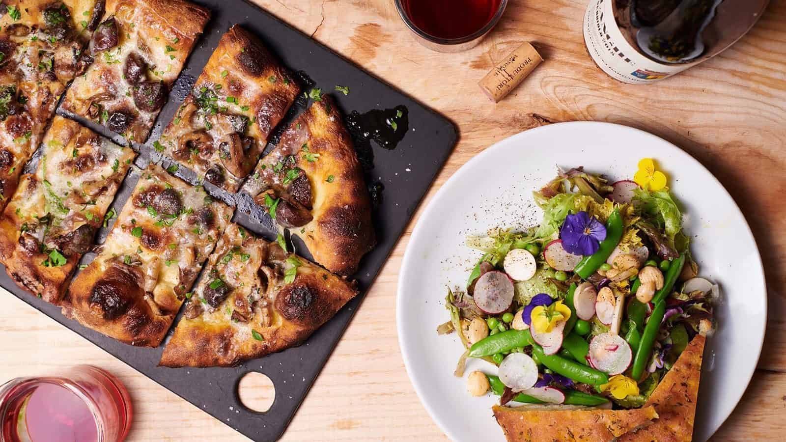 A wooden table hosts a delectable Seattle Pizza on a rectangular slate board, accompanied by a mixed salad on a white plate, a cork, and partially visible bottle and glass of red wine—a saucy serenade for the senses.