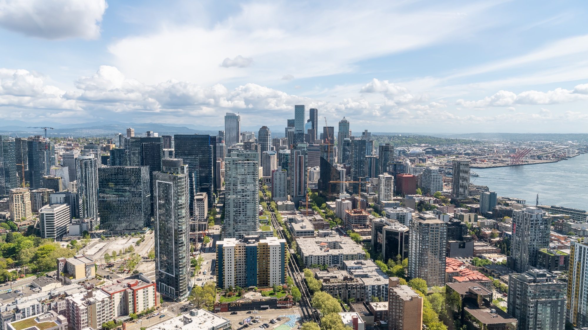 Aerial view of Seattle's skyline reveals tall buildings and a vibrant waterfront, with scattered clouds lingering under the blue sky. Below, shared living spaces thrive amidst vacant offices in the bustling cityscape.