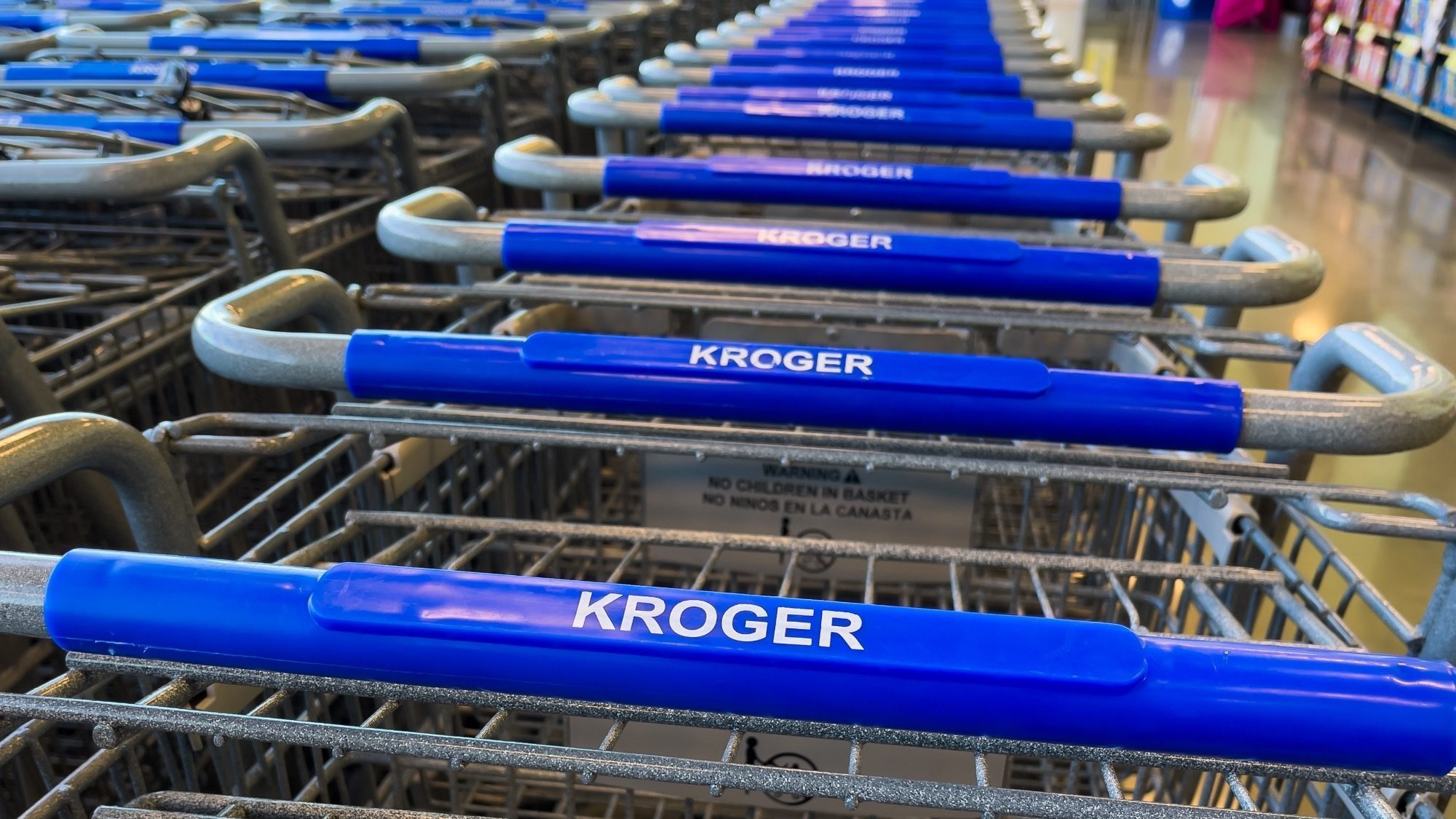 A row of shopping carts with blue handles labeled "Kroger" and hints of a strategic merger with Albertsons is lined up in a store.