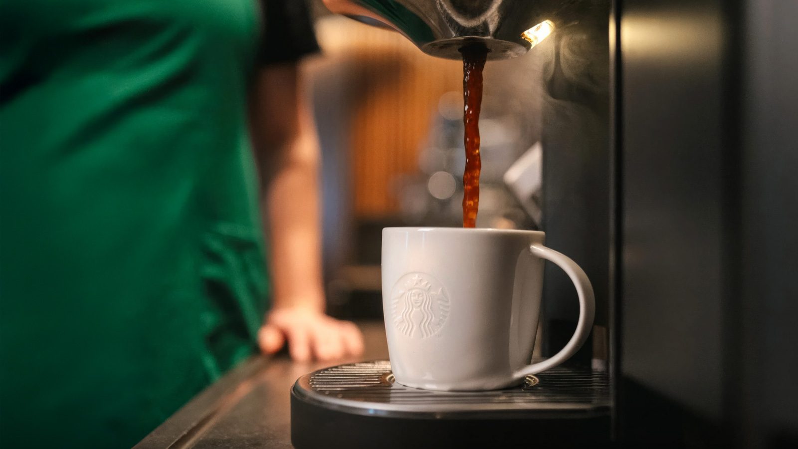 Aromatic coffee flows into a white ceramic mug adorned with a logo, fresh from the espresso machine. In the background, a person wearing a green apron—likely from Starbucks—attends to handwritten notes, adding a personalized touch to the bustling café scene.
