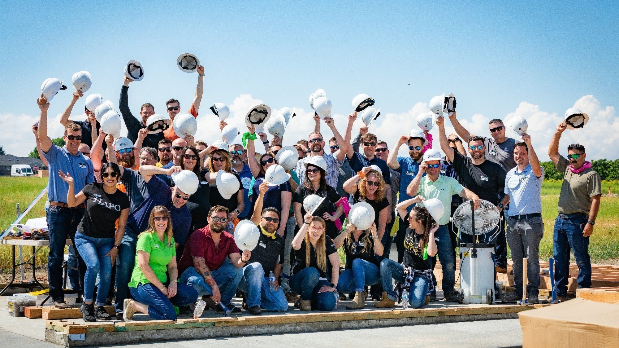 A group of construction workers cheering with hard hats in the air, standing together on a construction site under a clear blue sky, exemplifying community building.