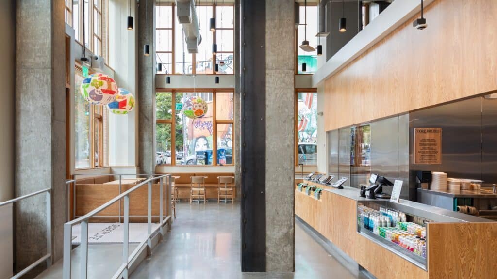 Modern cafe interior with tall ceilings, concrete columns, wooden accents, and colorful globe lights. Counter with self-service area, seated area to the left, and street view outside in bustling Capitol Hill. It's the perfect spot for enjoying a moment of calm before the lunch rush at Sweetgreen next door.